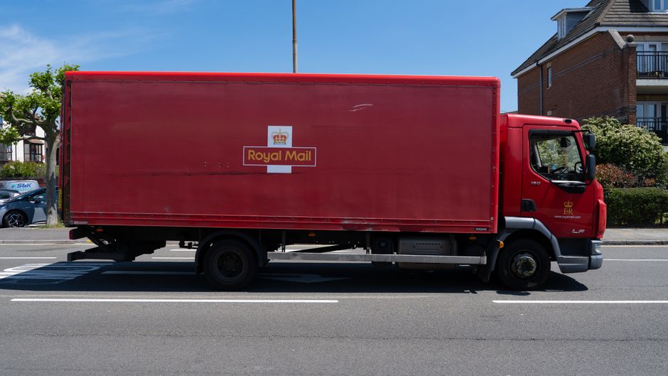 A red Royal Mail delivery truck parked on a residential street, with the side door partially open revealing the interior. The truck is positioned parallel to the curb, with the Royal Mail logo and branding visible on the side panel. The background shows a brick residential building with balconies and windows, and greenery including trees and bushes lining the street. The pavement is clear, and the truck appears to be involved in home relocation or furniture transport, possibly during a moving process managed by Edgware Man and Van, as indicated by the context of house removals and logistical support on the webpage about Barnet Council permit rules for house removals in Edgware.