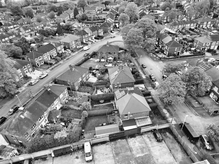 An aerial black-and-white photograph of a residential neighbourhood showing a variety of detached and semi-detached houses with pitched roofs, some featuring solar panels, and small front and back gardens. Several cars are parked along narrow roads and driveways, with one street curving through the area. The scene includes large trees, hedges, and fences separating properties. In the foreground, a flat rooftop with a small garage and parked vehicles is visible, indicating an urban setting typical of house removals or home relocation activities. The image captures the quiet, organized layout of houses and the surrounding environment, reflecting a typical suburban street scene where a professional moving service such as Edgware Man and Van might operate during furniture transport or packing and moving processes, possibly involving loading items onto a van parked nearby.