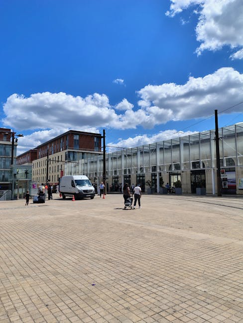 A spacious outdoor area outside the Broadwalk Centre in Edgware under a bright blue sky with scattered white clouds. The area features a large paved surface with rectangular concrete tiles. Several pe