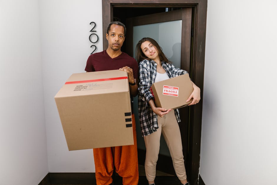 A man and a woman standing inside a doorway of a residential property, holding large cardboard boxes during a home relocation. The man, dressed in an orange pair of trousers and a maroon top, is holding a sizable cardboard box with red and black tape, while the woman, in beige trousers and a checked shirt, holds a smaller box marked with a 'Fragile' sticker. The background shows a white wall with the house number partially visible, and a dark door frame leading to another room. The environment indicates an active loading process involving packing materials and boxes, typical of furniture transport and packing and moving activities, supported by Edgware Man and Van’s removals services.