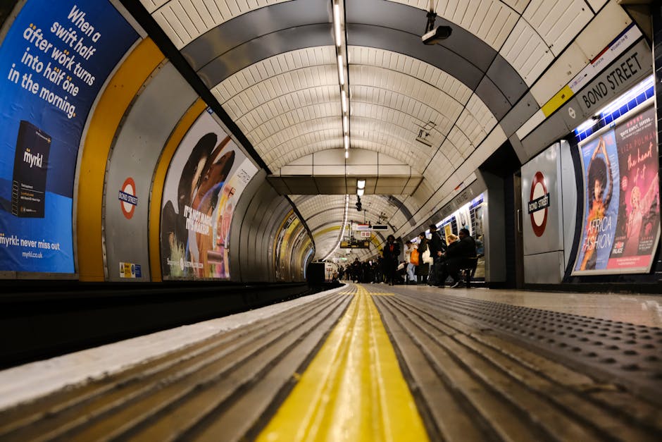 A low-angle view of the underground platform at Edgware Tube Station showing the curved, tiled ceiling with fluorescent lighting. Passengers are seated on benches along the platform edge, some standing and waiting for arriving trains. Opposite the platform, advertisements are displayed on the station walls, including promotional posters for products and events, with visible London Underground signage indicating the station name as 'BOND STREET.' The platform surface features tactile paving strips with a yellow line along the edge, and the station lighting creates a well-lit environment suitable for a home relocation or moving process. The image captures the typical environment where furniture transport, loading, and packing occur during house removals, with the overall scene emphasizing the importance of efficient logistics in moving services provided by companies like Edgware Man and Van.