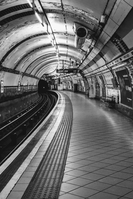A black and white photograph of an empty underground train station platform, featuring curved walls and ceiling with visible pipes and ventilation ducts. The platform is lined with tactile paving strips near the edge, and the floor is tiled with a diamond pattern. Indoor lighting illuminates the space, casting reflections on the shiny tiled surfaces. Advertising posters and informational signs are attached to the tiled walls along the platform. The station is equipped with safety features and electrical equipment, indicative of a typical urban transportation setting, suitable for house removals and furniture transport planning by Edgware Man and Van.