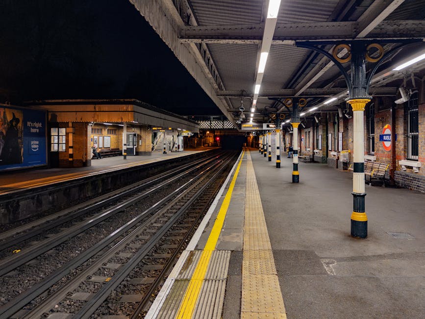 A low-angle view of the underground platform at Edgware Tube Station showing the curved, tiled ceiling with fluorescent lighting. Passengers are seated on benches along the platform edge, some standing and waiting for arriving trains. Opposite the platform, advertisements are displayed on the station walls, including promotional posters for products and events, with visible London Underground signage indicating the station name as 'BOND STREET.' The platform surface features tactile paving strips with a yellow line along the edge, and the station lighting creates a well-lit environment suitable for a home relocation or moving process. The image captures the typical environment where furniture transport, loading, and packing occur during house removals, with the overall scene emphasizing the importance of efficient logistics in moving services provided by companies like Edgware Man and Van.