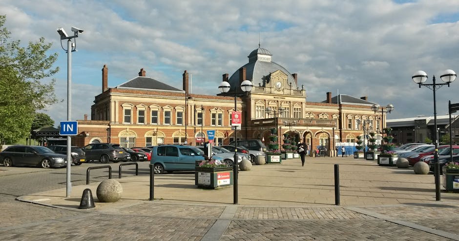 A large, historic train station building made of red brick and stone features multiple arched windows and a prominent central tower with decorative elements. In front of the station, there is a spacious paved area with several parked cars, including a light blue van and dark-colored vehicles, indicating a parking lot used for house removal and furniture transport services. The foreground includes black metal barriers, large spherical stone bollards, and flower planters, with a blue 'IN' parking sign visible on a pole. Bright overhead streetlights and vintage-style lamp posts line the area, while pedestrians walk nearby, suggesting a busy urban environment suitable for loading and unloading furniture during home relocation processes. The sky is partly cloudy, providing natural lighting that illuminates the scene clearly, reflecting typical city logistics for removals, packing, and moving activities. Edgware Man and Van’s presence is implied through the context of house removals and transport logistics in this setting.