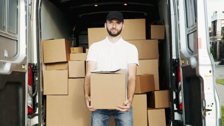 A man with a beard, wearing a white polo shirt and a black cap, is standing at the rear of an open moving van filled with neatly stacked cardboard boxes of various sizes, some wrapped in plastic. The boxes are arranged to facilitate efficient loading and safe transport during a home relocation, with some positioned on the pavement outside the van while others are inside. The man's expression is neutral as he holds a medium-sized box close to his chest, with a clipboard resting on top of it. The moving van, parked on a residential street visible in the background, features a spacious cargo area with a dark interior. This scene illustrates the packing and loading process involved in professional removals, with the focus on careful handling, organized storage, and transportation logistics. Edgware Man and Van offers services related to furniture transport and home moves, supporting efficient relocation in the local area.